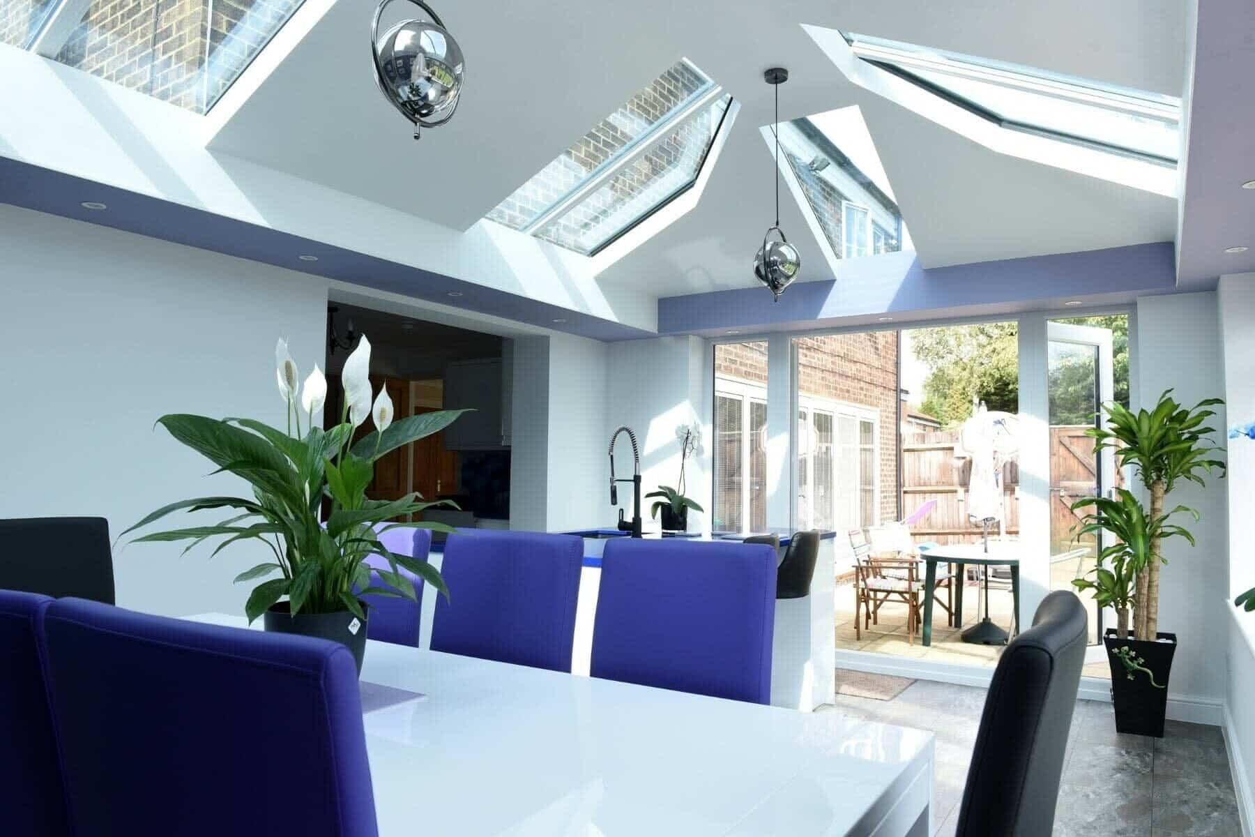 A modern dining area with a white table, purple and black chairs, indoor plants, and skylights. Large uPVC windows and glass doors let in sunlight, revealing an outdoor patio with a table and parasols in Horsham.