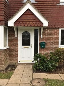 A white uPVC front door with a decorative glass panel and brass letterbox is set in a red brick house in West Sussex. There’s a green postbox on the wall, a bush by the door, and a tiled path leading to the entrance.