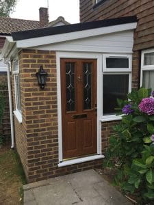 A brick porch extension with a composite front door, decorative glass panels, uPVC white-framed windows, a black wall lantern, and purple hydrangea flowers growing nearby in West Sussex.