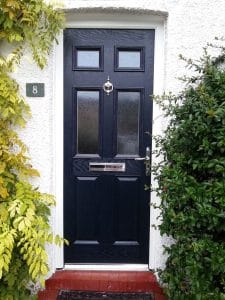 A black composite front door with a letterbox and door knocker, set in a white textured wall. Green and yellow leafy plants frame both sides of this West Sussex entrance.