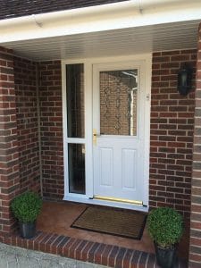 A white composite front door with decorative glass panels, set in a red brick porch in West Sussex. There is a brown doormat in front and two potted green shrubs on either side. A wall-mounted light is on the right.