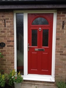 A bright red composite front door with a white frame and silver handle, knocker, and letterbox. The door is set in a brick wall with a textured glass panel on the left and flowerpots nearby in West Sussex.