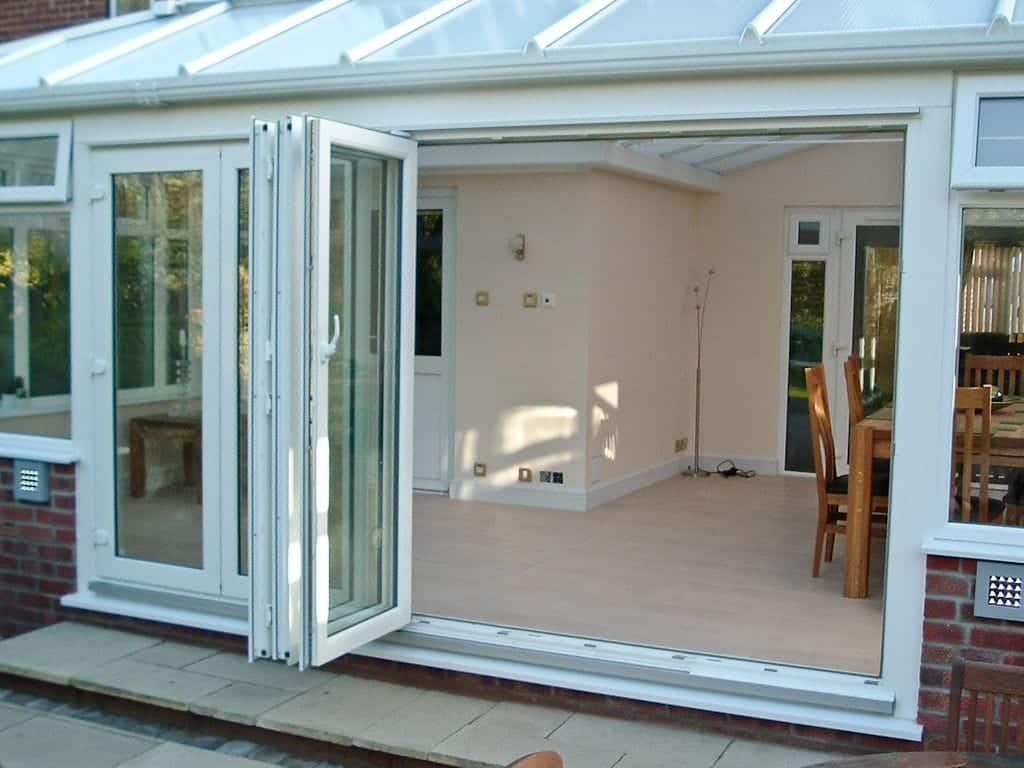 Open white uPVC folding glass doors lead into a bright, tiled conservatory with a dining table and chairs. The space has cream walls, large windows, and a partially visible door at the back - perfect for any West Sussex home.