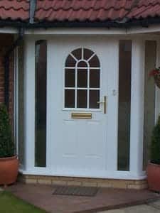 A white composite front door with a semi-circular window design and gold handle, flanked by narrow glass panels and two potted shrubs, set under a red tiled roof - perfect for welcoming guests in West Sussex.