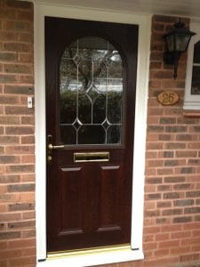 A brown composite front door with a decorative glass panel, brass handle, and letterbox, set in a brick wall with a house number plaque and a black outdoor lantern above, offering charming kerb appeal in West Sussex.