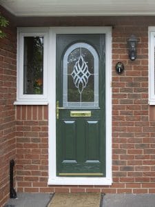 A green composite front door with a decorative glass panel, gold letterbox, and handle, set in a brick wall. A house number plaque and lantern-style light sit to the right - perfect for charming West Sussex homes.