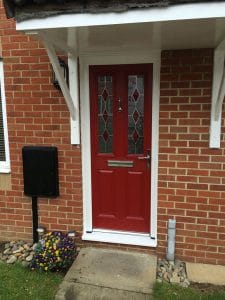A striking red uPVC front door with glass panels set in a brick wall, featuring a letterbox, door knocker, and handle. A black postbox, colourful flower bed, and concrete path complete this charming West Sussex entrance.