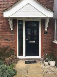 A black composite front door with a letterbox, set in a red brick wall with white trim, two side windows, and a small porch roof. Plants and a welcome mat line the stone path of this charming West Sussex home.