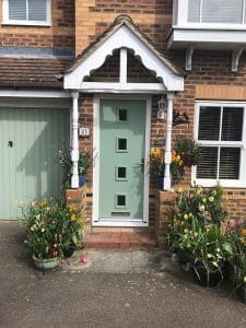 A light green composite front door with four square windows is centred between two white columns and flowering potted plants. The brick house, found in West Sussex, features a garage with a matching green door and white-trimmed windows.