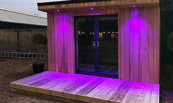 A small wooden garden office or shed with a deck, featuring uPVC-framed glass double doors illuminated by purple LED lights at dusk - perfect for stylish workspaces in Crawley.