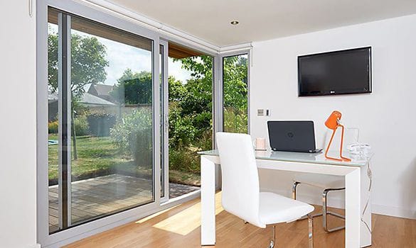 A modern home office with a white desk, white chair, orange lamp, laptop, and glass of water. Large uPVC glass doors and windows reveal a green garden outside in Billingshurst. A wall-mounted TV is above the desk.