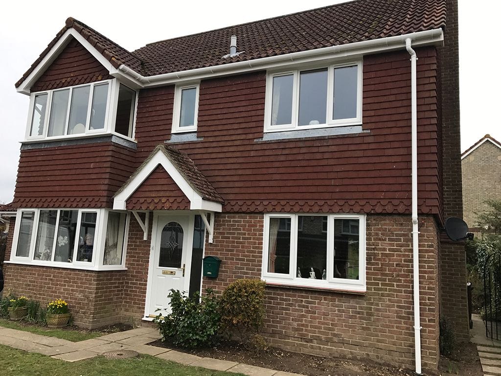 A two-storey house in West Sussex with a brown tiled roof, red wooden cladding on the upper floor, and brown brick on the lower. White uPVC-framed windows and a white front door with a small porch overlook a charming garden.