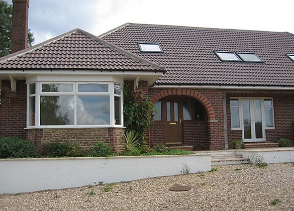 A single-storey brick house in Horsham with a tiled roof, large bay window, and arched wooden front door. Several rooflights and modern uPVC features enhance the roof, while a gravel drive welcomes you at the front.