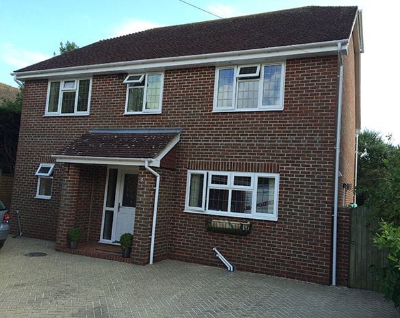 A two-storey red brick house in Horsham with a tiled roof, uPVC white-framed windows, a small covered porch, and a paved drive. Potted plants sit by the entrance, and a wooden fence stands to the right.