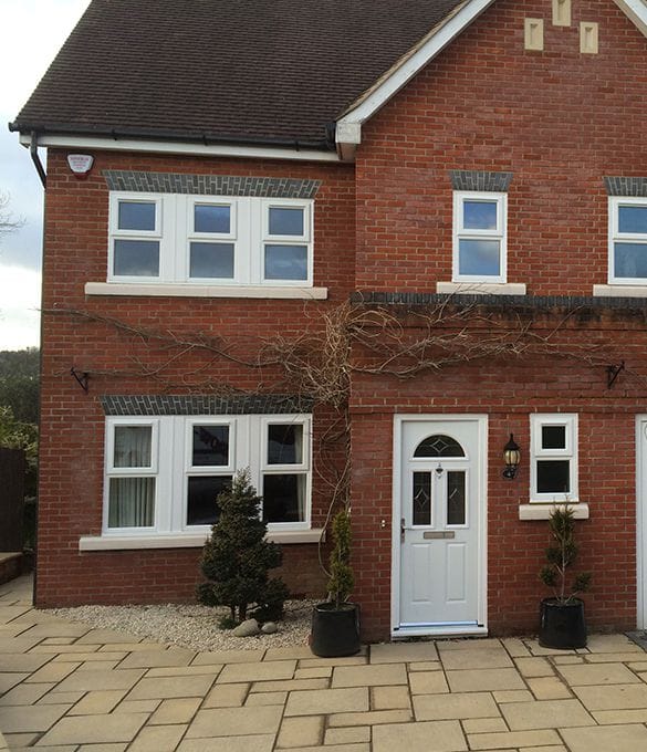 A red-brick house in Horsham with white uPVC-framed windows, a white front door, and potted plants by the entrance. The house features a paved drive and a small landscaped area with shrubs.