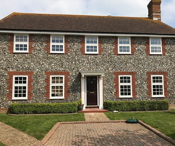 upvc windows in a two-storey stone house in Horsham with a dark tiled roof, red brick accents around the uPVC windows, a dark composite front door with columns, trimmed hedges, a paved path, and a grassy lawn.