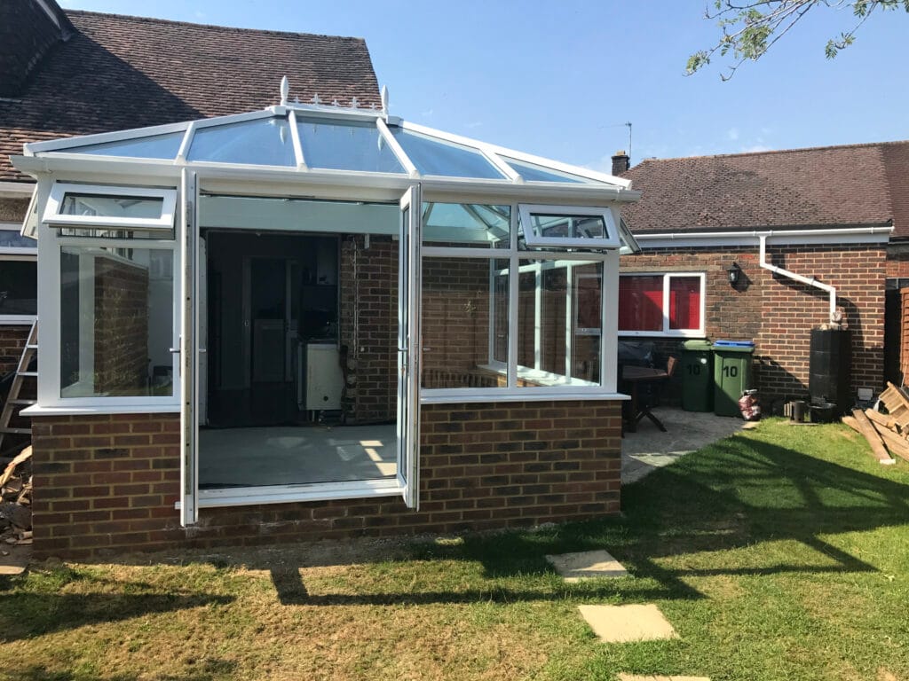 A uPVC glass conservatory with open double doors is attached to a brick house in Horsham. The conservatory has a glass roof and windows, and the back garden includes green grass, paving stones, bins, and a wooden bench.